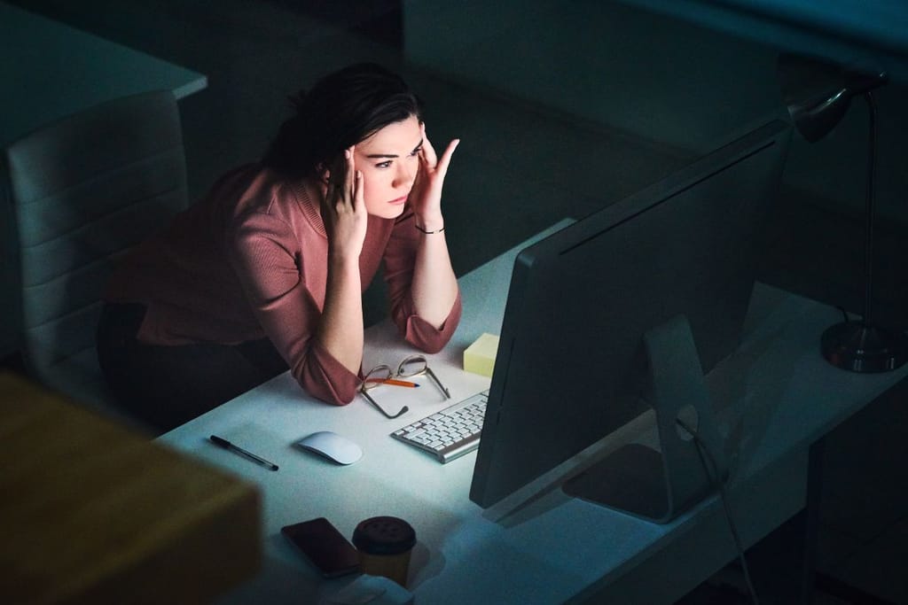 Nainen istuu myöhään illalla koneella ja tuijottaa näyttöä sormet ohimoilla. Hän on selkeästi stressaantunut.