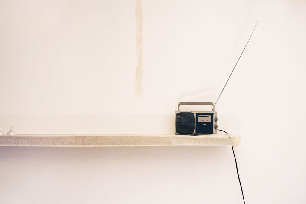 Pienikokoinen radio seisoo valkoisella hyllyllä vaaleaa seinää vasten. Radiosta nousee pitkä antenni, ja sen johto roikkuu hyllyn reunalta alaspäin.