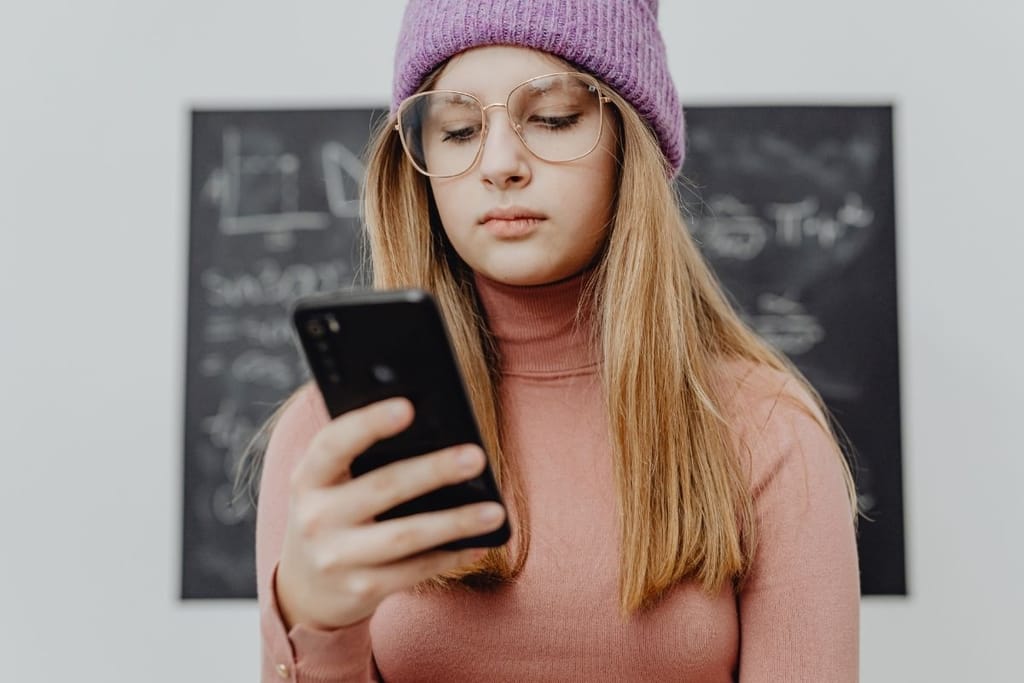 Teini-ikäinen tyttö selaa puhelinta violetti pipo päässä. Tausta on valkoinen ja selän takana on liitutaulu, jolla on 