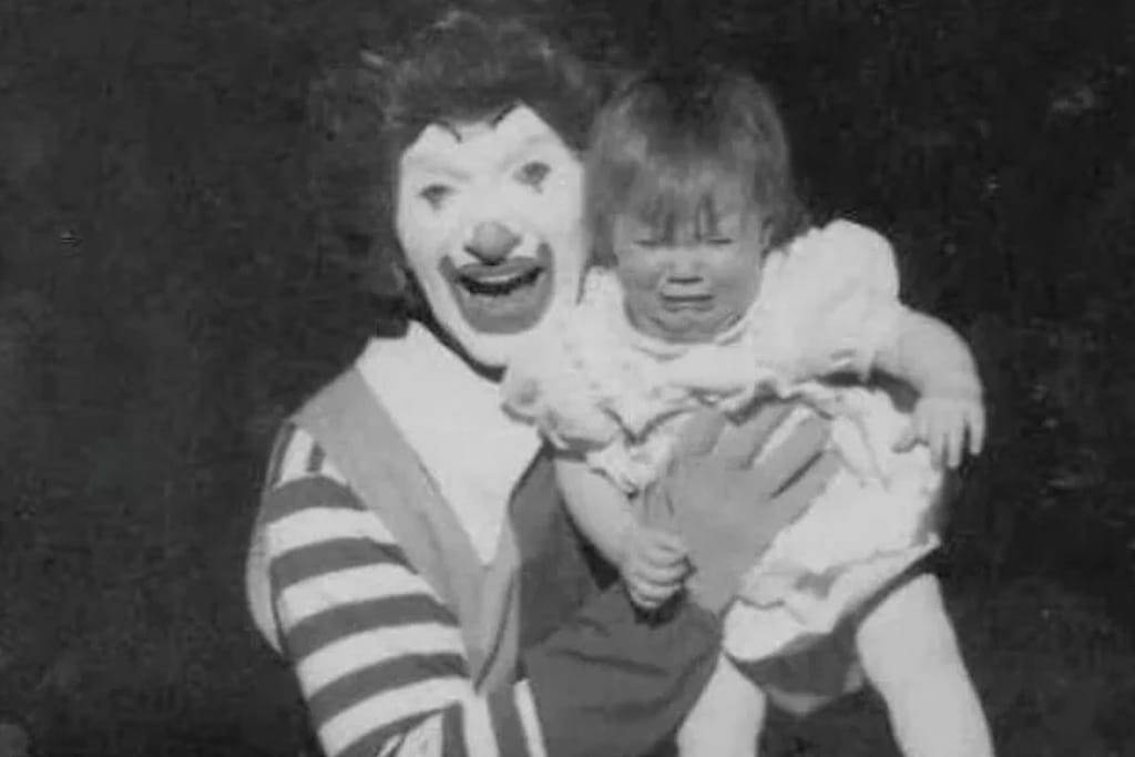 Vintagekuva varhaisesta Ronald McDonald -hampurilaisketjun maskotiksi pukeutuneesta näyttelijästä, joka pitelee itkevää vauvaa sylissään mustaa taustaa vasten.