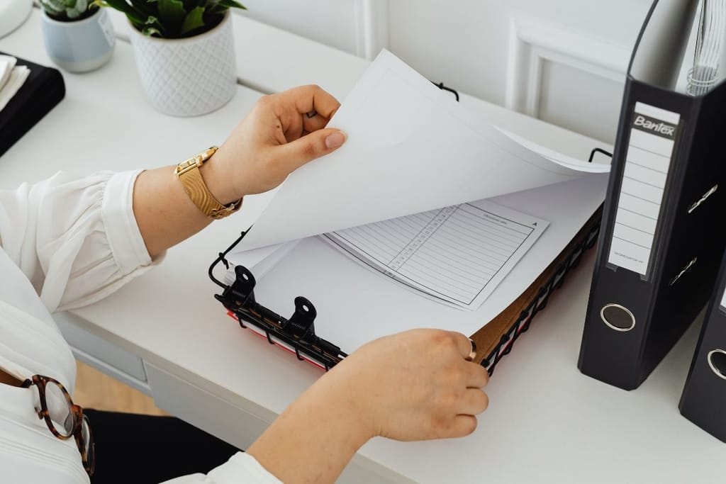 Naisen kädet selailevat yrityksen lomakkeita, joissa on sarakkeita. Naisella on kultakello, silmälasit roikkuvat paidankauluksesta ja taustalla on mustia kansioita. 