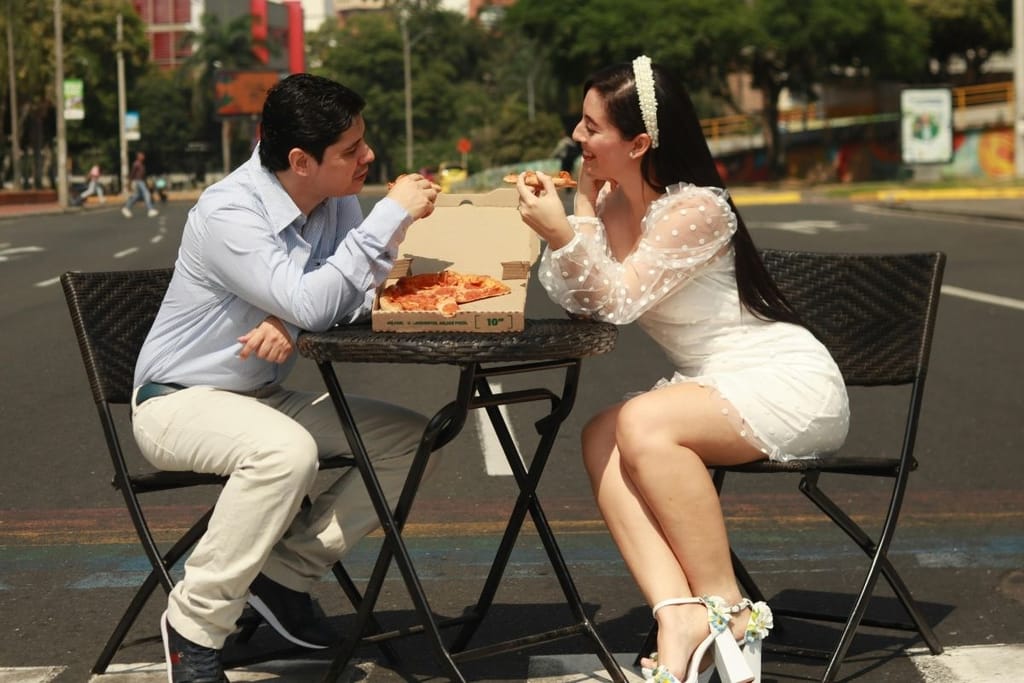 Kaksi ihmistä istuu ulkona pienessä pöydässä ja jakaa pizzapalan rennolla treffeillä kaupunkiympäristössä.