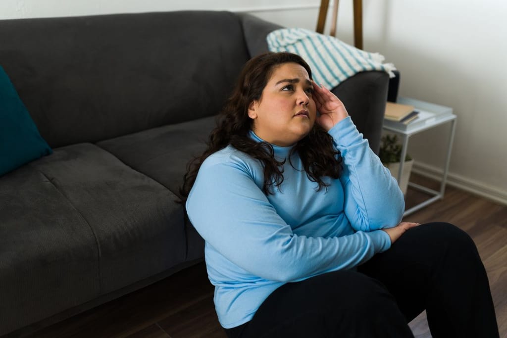 Pluskokoinen nainen istuu lattialla sohvan vieressä ja nojaa päätään käteensä olohuoneessa.