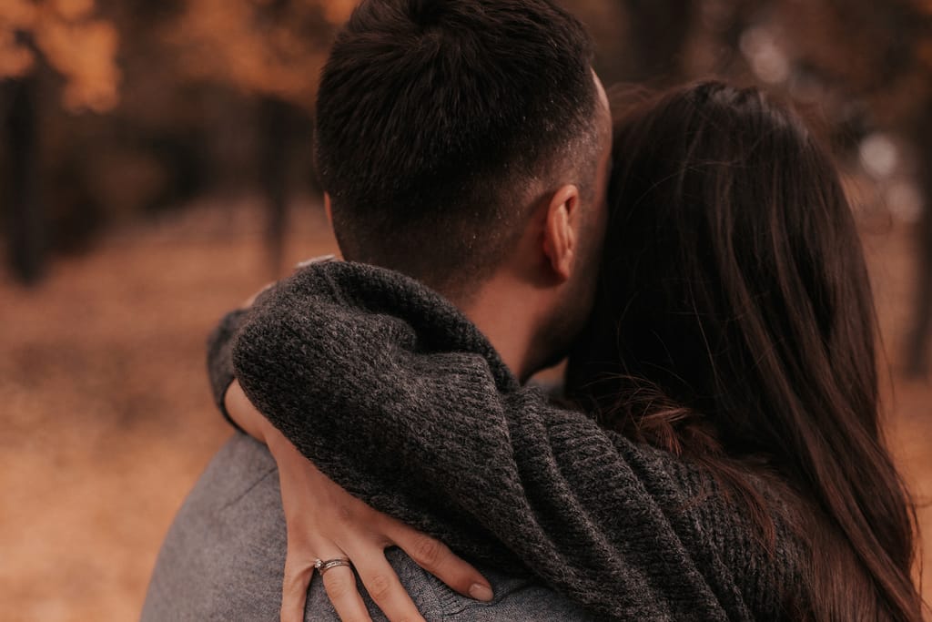 Pariskunta halaamassa ulkona syksyisessä puistossa, kuvattuna takaapäin; toisen käsi lepää kumppanin niskalla ja sormessa näkyy sormus.