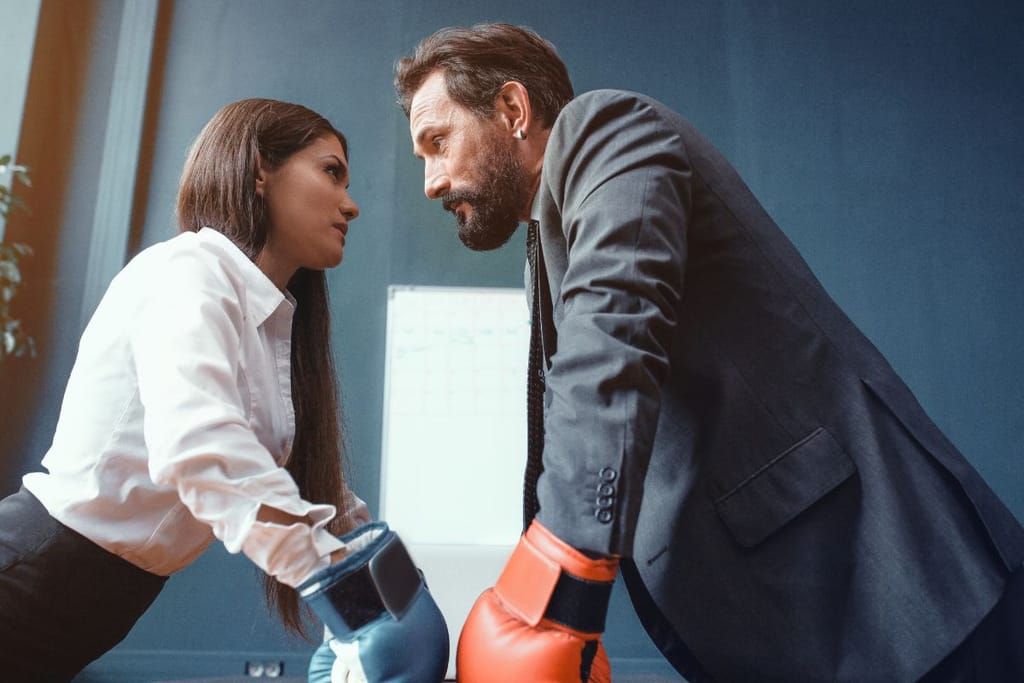 Kaksi ihmistä seisoo vastakkain toimistossa nyrkkeilyhanskat kädessä, katseet lukittuneina – symboloi riitaa tai valta-asetelman kamppailua.