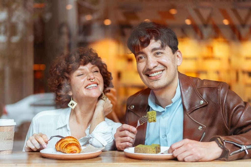 Kaksi nuorta istuu kahvilan ikkunan vastaisilla penkeillä ja näyttää iloisilta. He ovat syömässä. Mies hymyilee haarukka kädessään ja nainen on silmät kiinni. He ovat kahvilaympäristössä, jossa on lämmin valaistus.