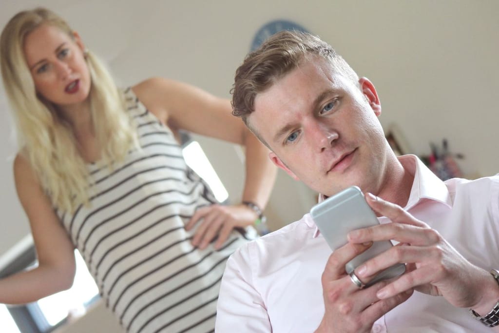 Mies selaa puhelintaan, nainen seisoo taustalla kädet lanteilla epäilevän näköisenä.