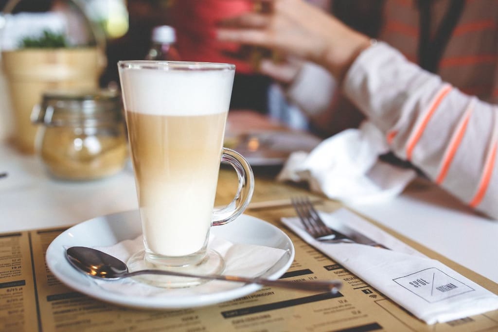 Lasiin kaadettu latte macchiato valkoisella lautasella kahvilapöydällä, taustalla henkilö nauttimassa juomaa.