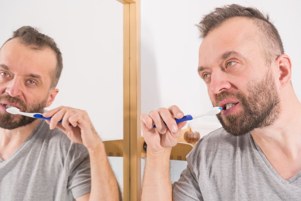 Partainen mies harjaa hampaitaan peilin edessä kylpyhuoneessa. Hänen heijastuksensa näkyy peilissä, ja hän katsoo keskittyneenä omaa kuvaansa.
