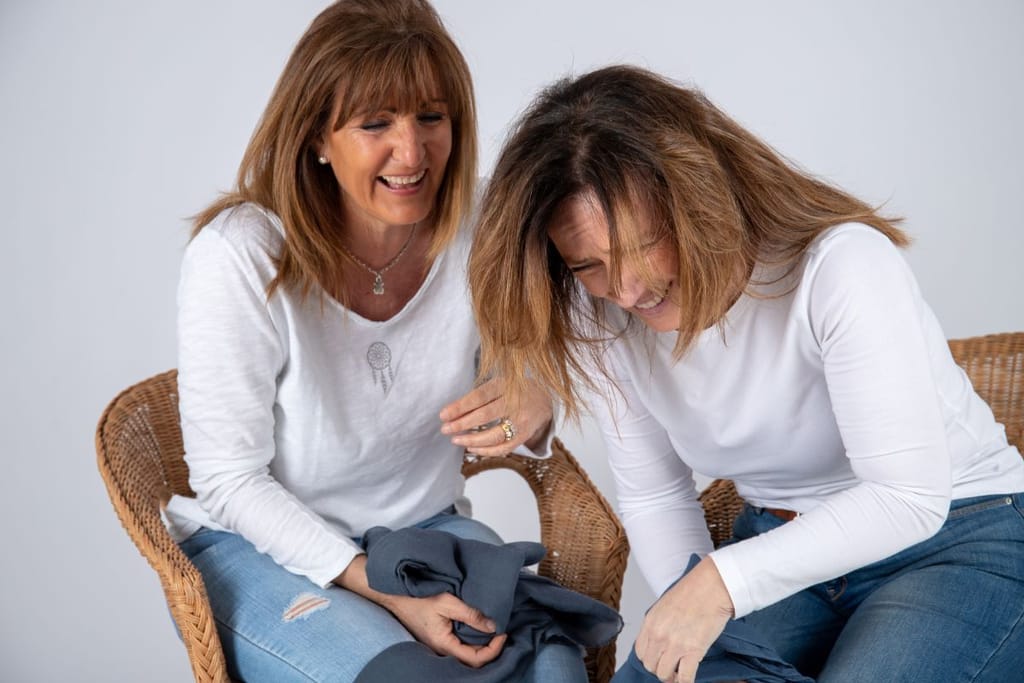 Kaksi aikuista naista istuu vierekkäin ja nauraa. Molemmilla on vaaleat paidat ja farkut. 