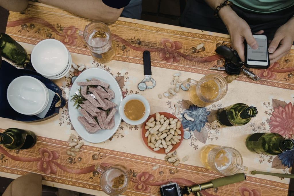 Ystäväporukka istuu pöydän ääressä oluiden ja naposteltavien kanssa. Yksi katsoo puhelintaan muiden seurassa. Kuva havainnollistaa, miten puhelin voi viedä huomion pois yhteisestä hetkestä.