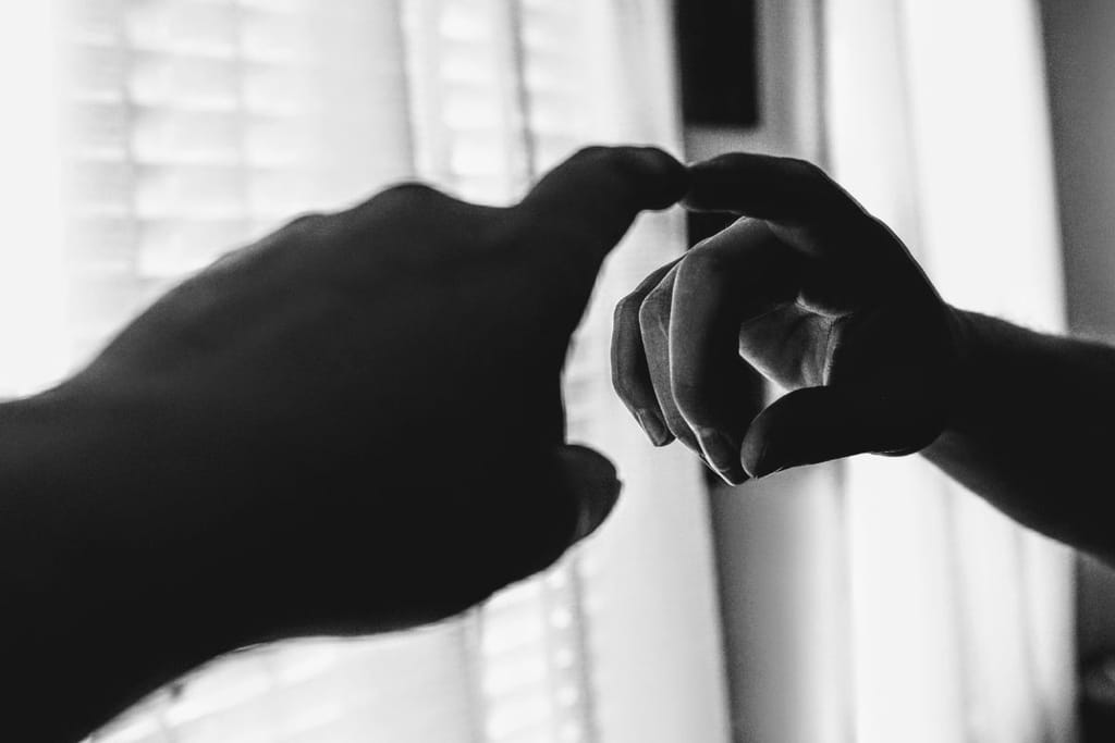 Kaksi kättä koskettaa toisiaan peilikuvan kautta mustavalkoisessa kuvassa, taustalla näkyy ikkunan valo. 