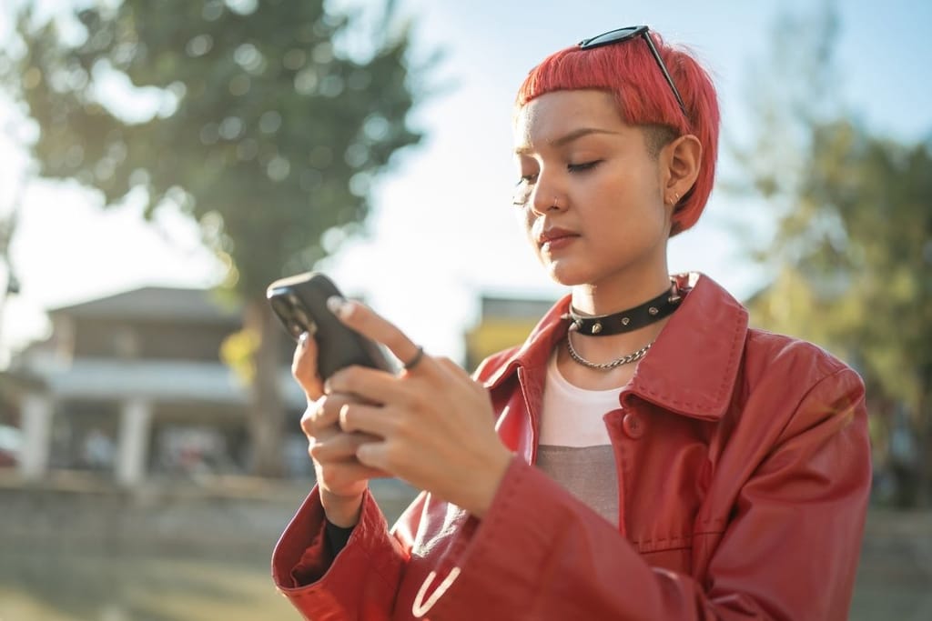 Lyhyttukkainen nuori seisoo ulkona ja katsoo puhelinta kädessään. 