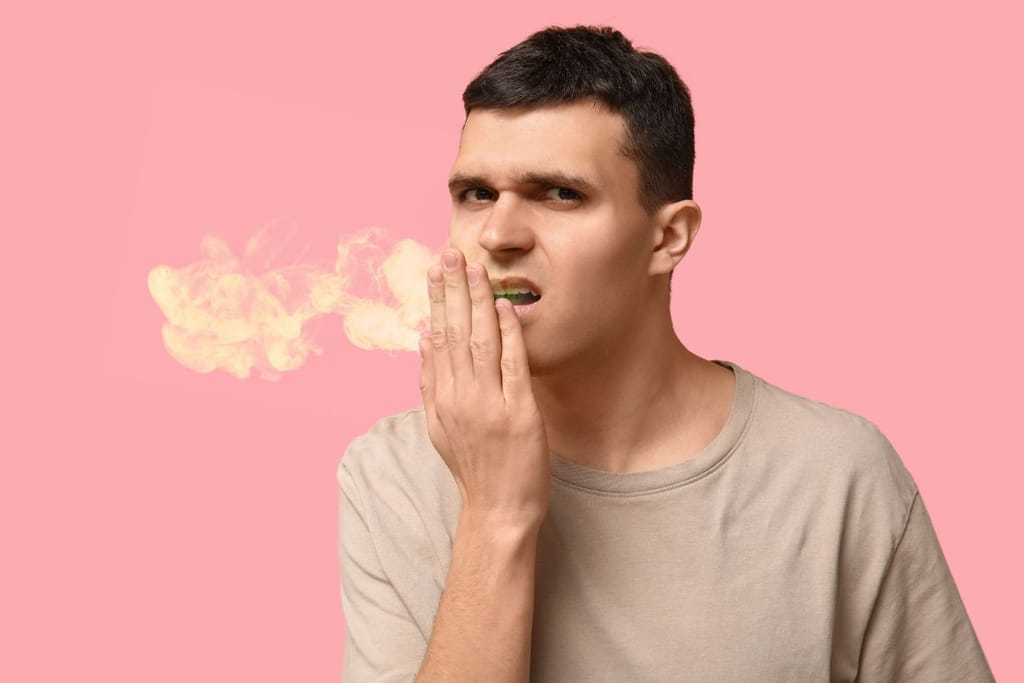Nuorehko mies vaaleassa paidassa pitää kättään suunsa edessä ja katsoo kameraan. Hänen suustaan leijailee kellertävää savua, joka kuvastaa pahanhajuista hengitystä, vaaleanpunaisella taustalla.