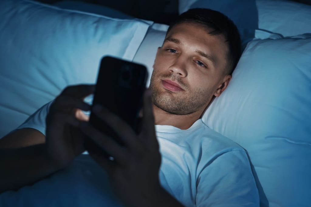 Mies makaa sängyssä pimeässä huoneessa ja katsoo puhelintaan.
