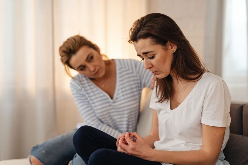 Kaksi naista istuu sohvalla, toinen lohduttaa toista koskettamalla olkapäätä, tunnelma on vakava.