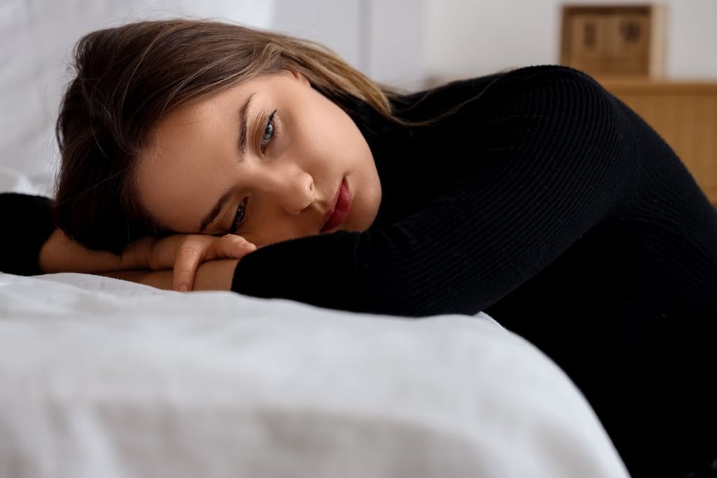 Nainen näyttää surulliselta ja hylätyltä. Hän nojaa valkeaa sängynreunaa vasten ja katselee kaukaisuuteen ajatuksissaan.