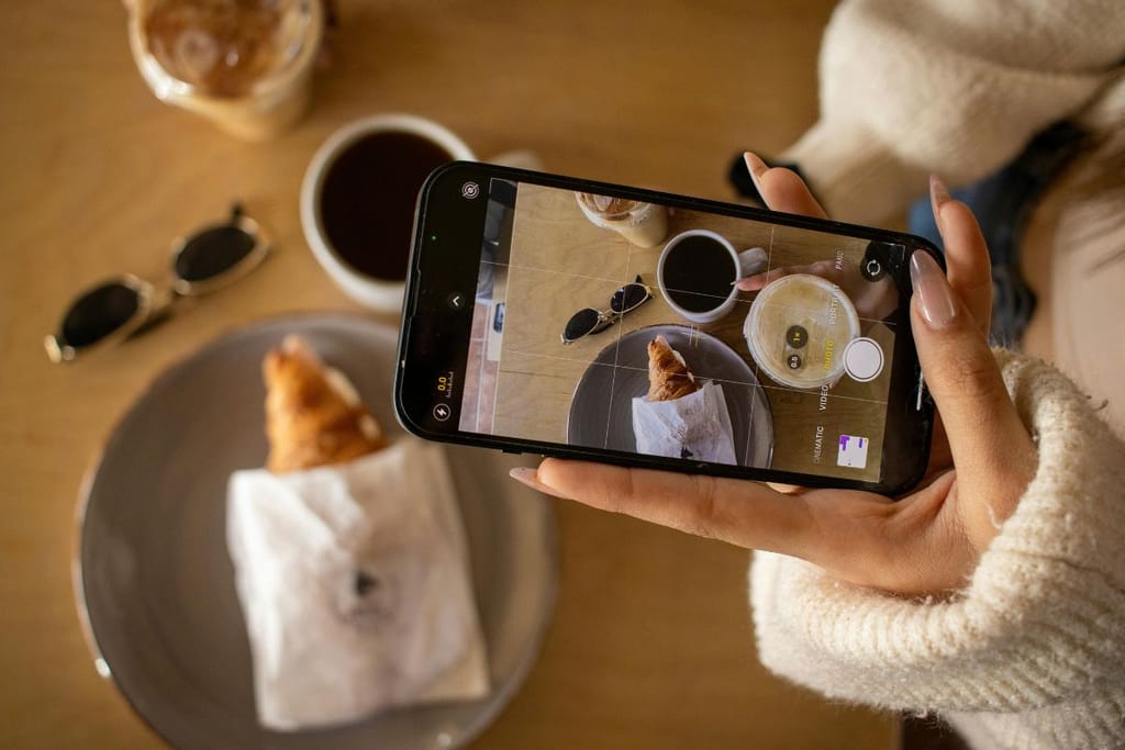 Nainen kuvaa älypuhelimella kahvilatuotteita, kuten paperiin käärittyä croissanttia ja kahvikuppia.