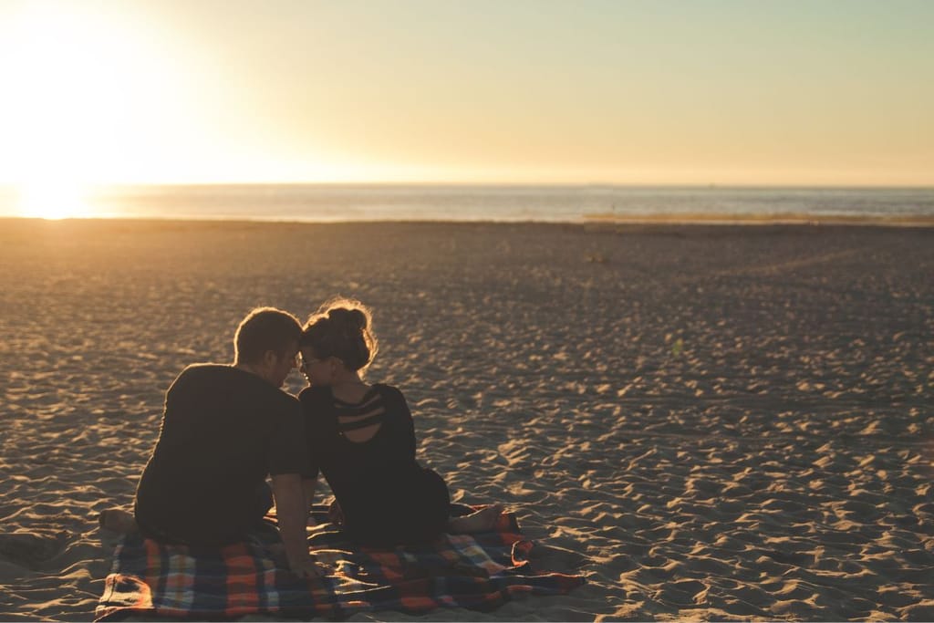 Nuori pari istuu autiolla hiekkarannalla vierekkäin otsat vastakkain. On auringonlasku ja he ovat selkeästi pariskunta, joka istuu yhdessä viltillä. 