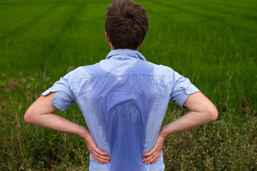 Mies seisoo ruohokentän laidalla kädet lanteilla ja hänen paitansa on selkäpuolelta läpimärkä hiestä. 