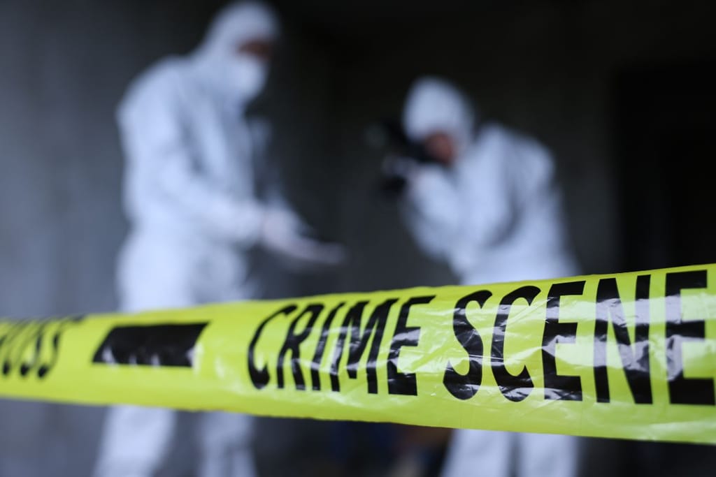 Kaksi valkoiseen suojapukuun pukeutunutta tutkijaa seisoo keltaisten poliisinauhojen takana mustaa taustaa vasten.