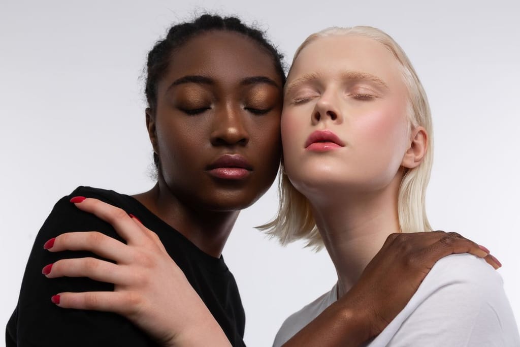 Kaksi naista eri ihonväreissä seisoo lähekkäin studioympäristössä. Molemmilla on silmät kiinni ja toisen käsi lepää toisen olkapäällä.