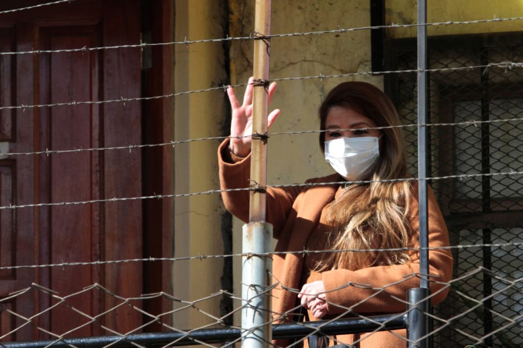 Nainen vilkuttaa piikkilanka-aidan yli alas. Hänellä on kasvoillaan valkoinen maski.