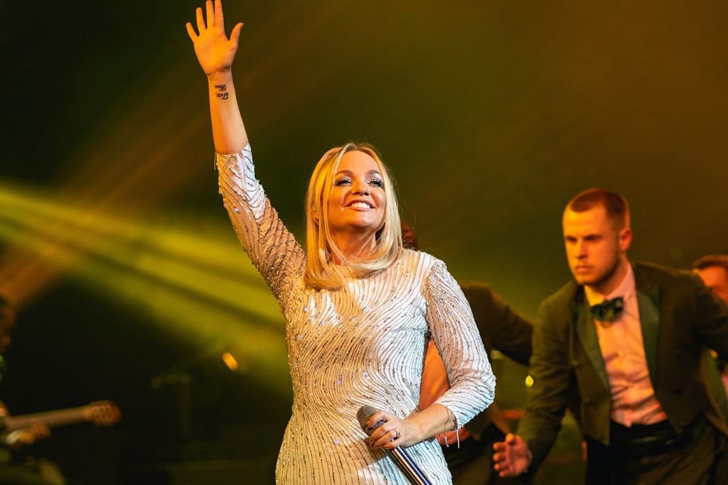 Vaaleahiuksinen naisartisti puristaa kädessään mikkiä ja on toinen käsi kohotettuna kattoon. Hän katsoo yleisöön ja hymyilee vaalea mekko yllään. Taustalla on mies tummassa puvussa.