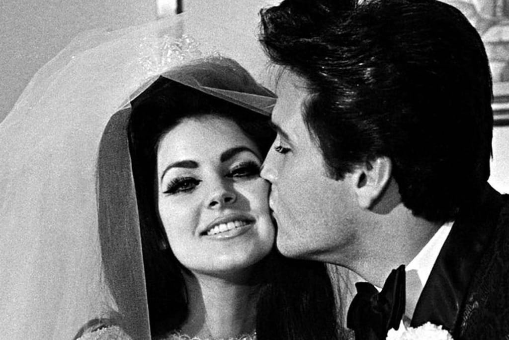 Elvis Presleyn ja Priscillan häissä otettu kuuluisa pusukuva. Lisa Marie katsoo kameraan morsiushuntu päässään ja hymyilee samalla kun Elvis antaa hänelle suukon poskelle. Mustavalkokuva. 