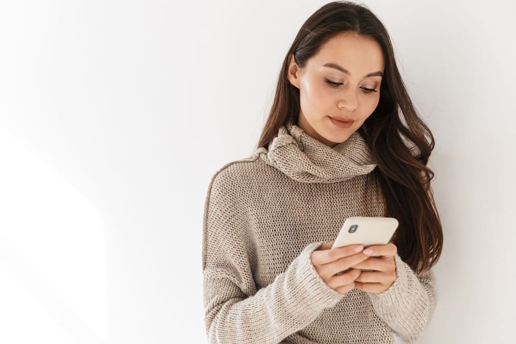 Nainen seisoo puhelimella beigen värisessä paidassa valkoista taustaa vasten. Hän näyttää iloiselta.