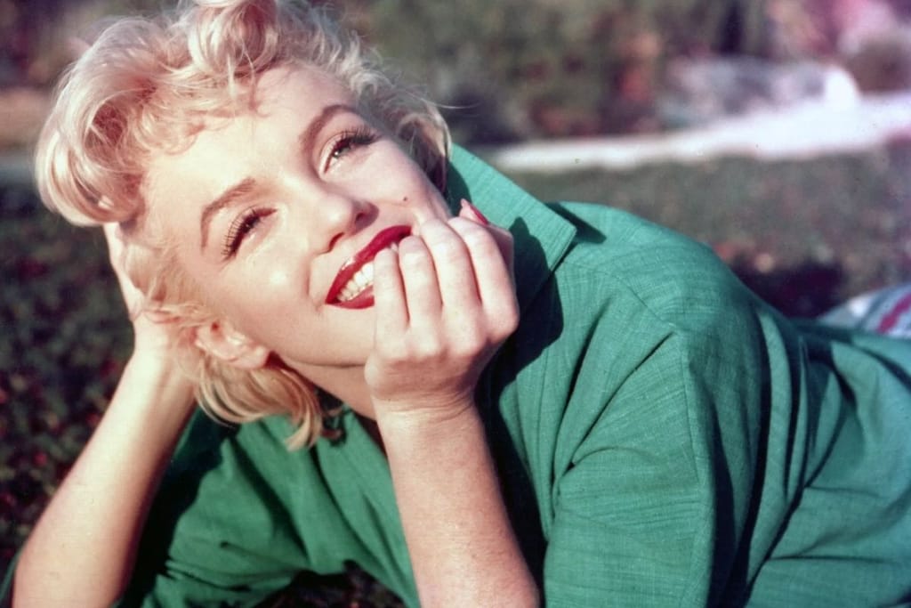 Marilyn Monroe vihreässä takissa makaa maassa ja hymyilee kameran ohi. Hänellä on unenomainen ilme. 