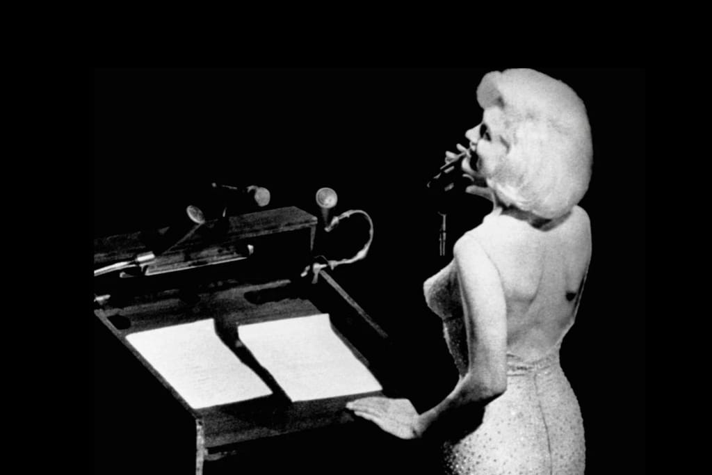 Marilyn Monroe esiintyy lavalla mikrofonin ääressä mustavalkoisessa kuvassa. Hän seisoo puhujapöntön vieressä yllään vartalonmyötäinen, paljettimekko ja laulaa yleisölle, kuvattuna sivulta tummalla taustalla.