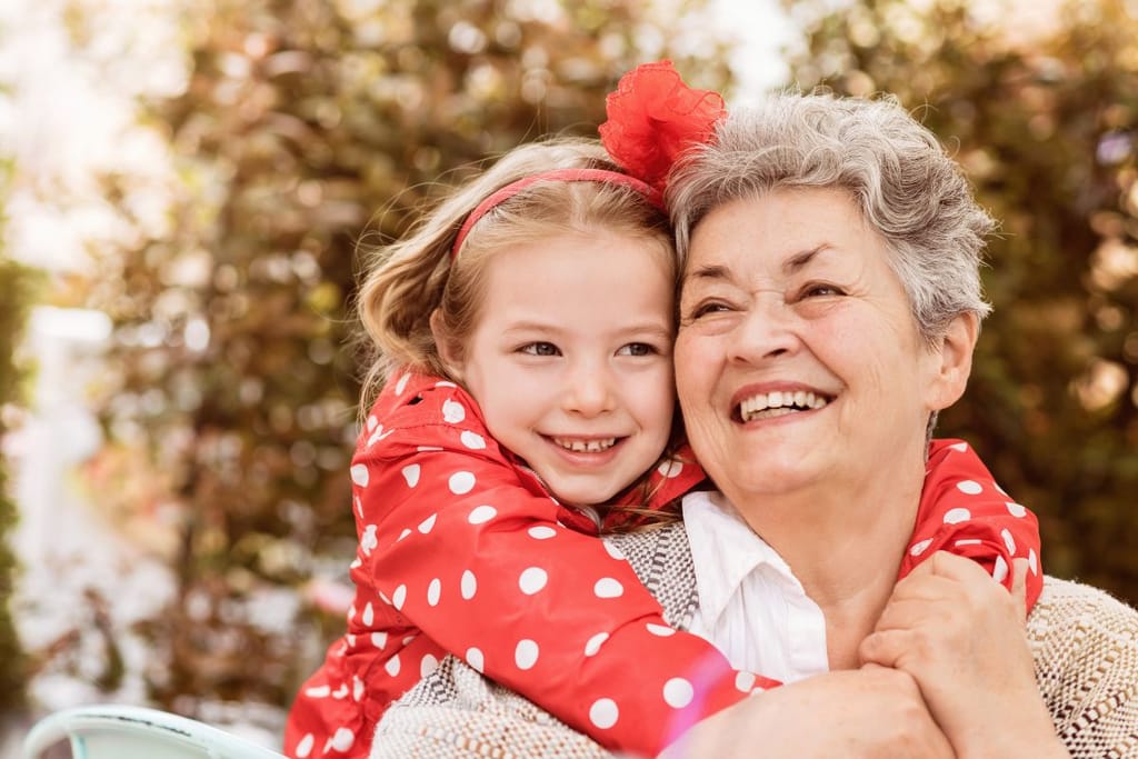 Iäkäs nainen ja pieni lapsi halaavat toisiaan ulkona, molemmat hymyilevät ja katsovat sivulle syksyisessä ympäristössä