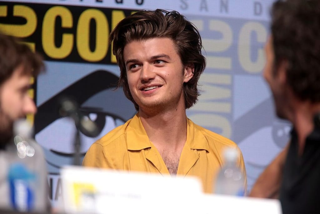 Ruskeahiuksinen Joe Keery istuu paneelikeskustelussa ja hymyilee muiden osallistujien välissä tapahtumaympäristössä.
