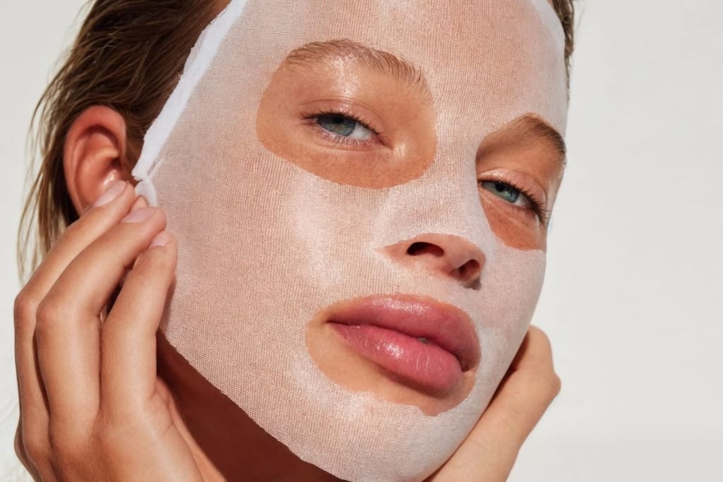 Naisella on kasvoillaan läpikuultava kasvonaamio, ja hän katsoo kameraan pitäessään sormiaan leukaperillä.