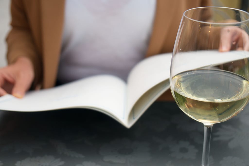 Henkilö lukee kirjaa pöydän ääressä, etualalla lasi valkoviiniä.