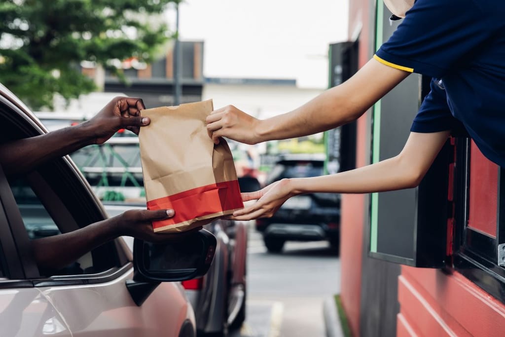 McDonaldsin ravintolan autokaistan luukulla ruoka vaihtaa omistajaa. Myyjä ojentaa paperisen pussin autosta kurottaville käsille. 