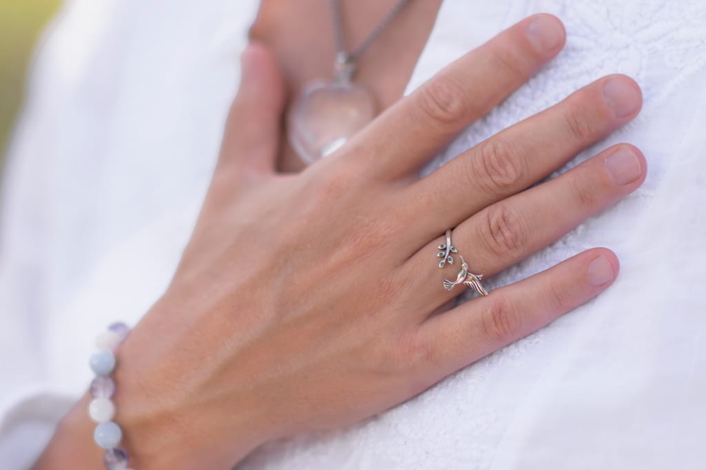 Lähikuva kädestä, jossa on sormus ja rannekoru, lepäämässä rinnan päällä valkoista vaatetta vasten.