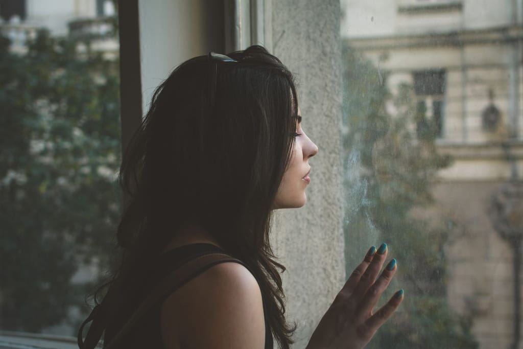 Nainen seisoo ikkunan edessä silmät kiinni ja pitää sormiaan ikkunaa vasten. Ikkunasta näkyy urbaani kaupunkimiljöö.