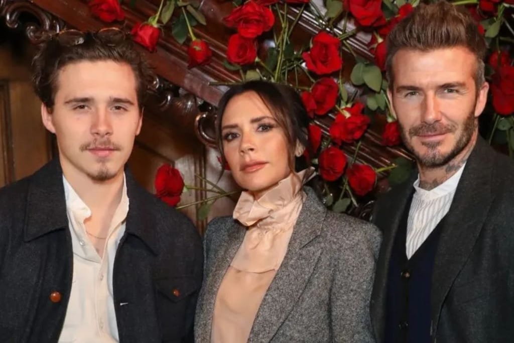 Kolme aikuista eli Brooklyn, Victoria ja David Beckham seisovat vierekkäin sisätilassa, taustalla runsaasti punaisia ruusuja ja koristeellinen seinäpinta.