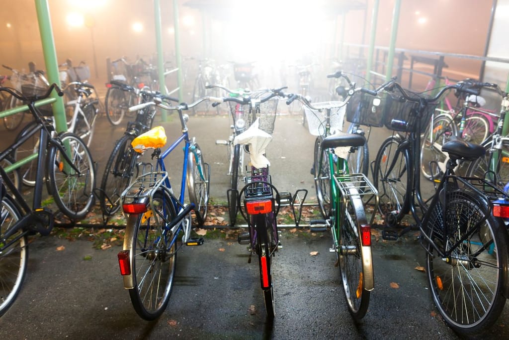 Polkupyöriä telineessä. Taustalla on kerrostalolähiön näköinen ympäristö, jossa on sumuinen sadekeli ja asffaltti on märkä ja täynnä ruskeita lehtiä. 