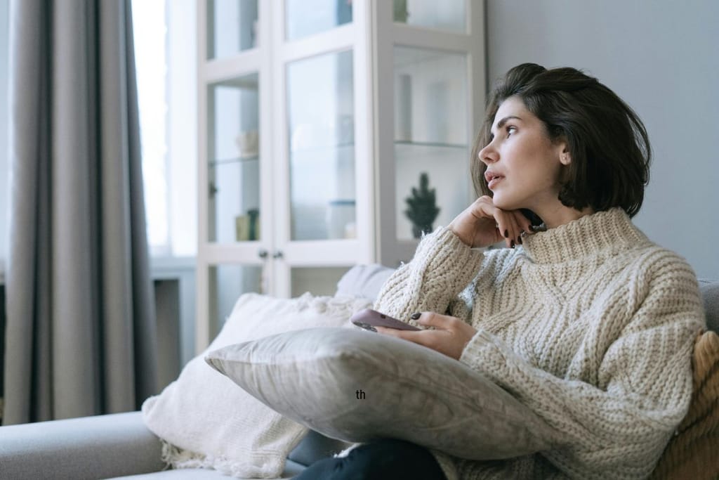 Aikuinen nainen istuu sohvalla sisätiloissa ja katsoo sivulle, kädessään älypuhelin.