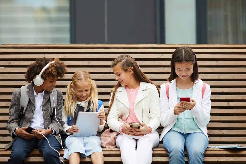 Neljä alakouluikäistä lasta istuu ulkona penkillä ja käyttää digilaitteita: älypuhelimia ja tablettia.