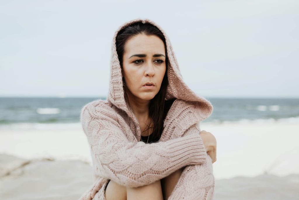 Nainen istuu neulehupparissa rannalla, ja hänellä on surullinen ilme. Taustalla on rantaviiva. 