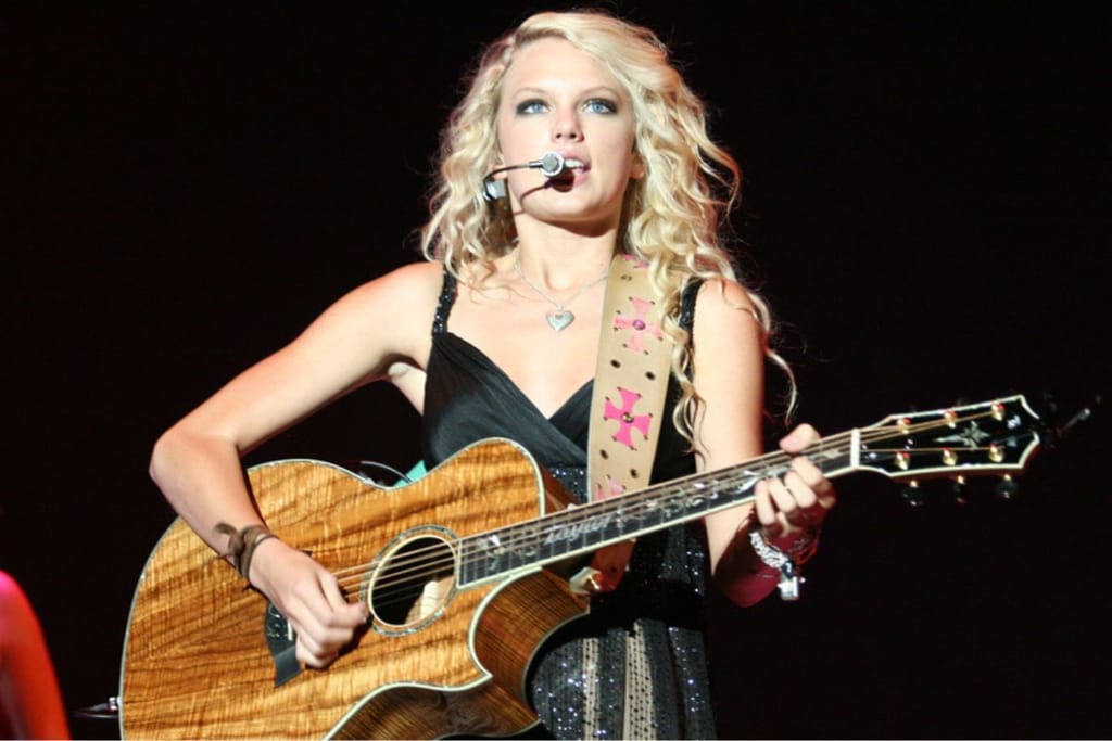 Taylor Swift esiintyy lavalla akustisen kitaran kanssa ja laulaa mikrofoniin konserttivalojen loisteessa tummaa taustaa vasten.