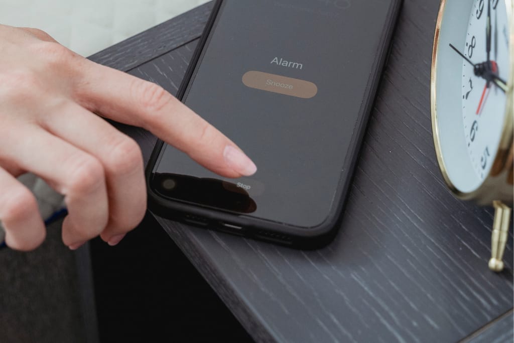 Sormi koskettaa älypuhelimen herätyskellon näytöllä olevaa “Stop”-painiketta yöpöydällä; vieressä näkyy perinteinen herätyskello.