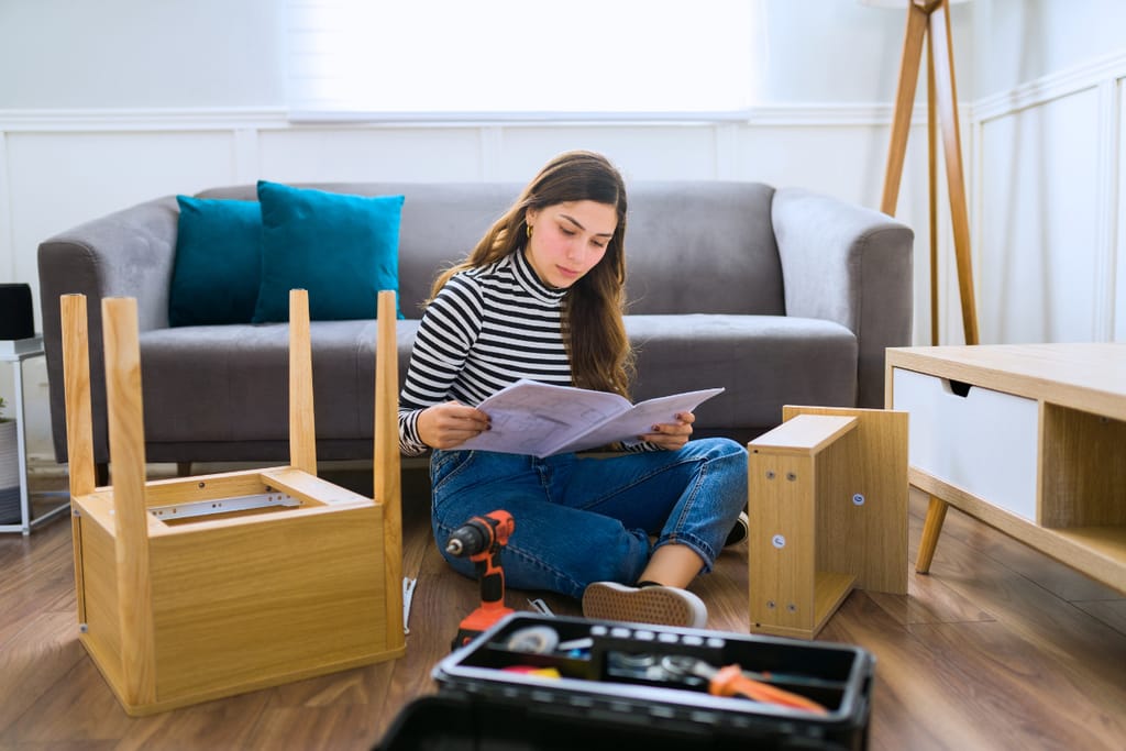 Nuori nainen istuu lattialla olohuoneessa ja lukee huonekaluohjeita ennen kalusteen kokoamista. Hänen ympärillään on puuosia ja työkaluja, jotka kertovat itsenäisestä arjen projektista ja aikuisen vastuista.
