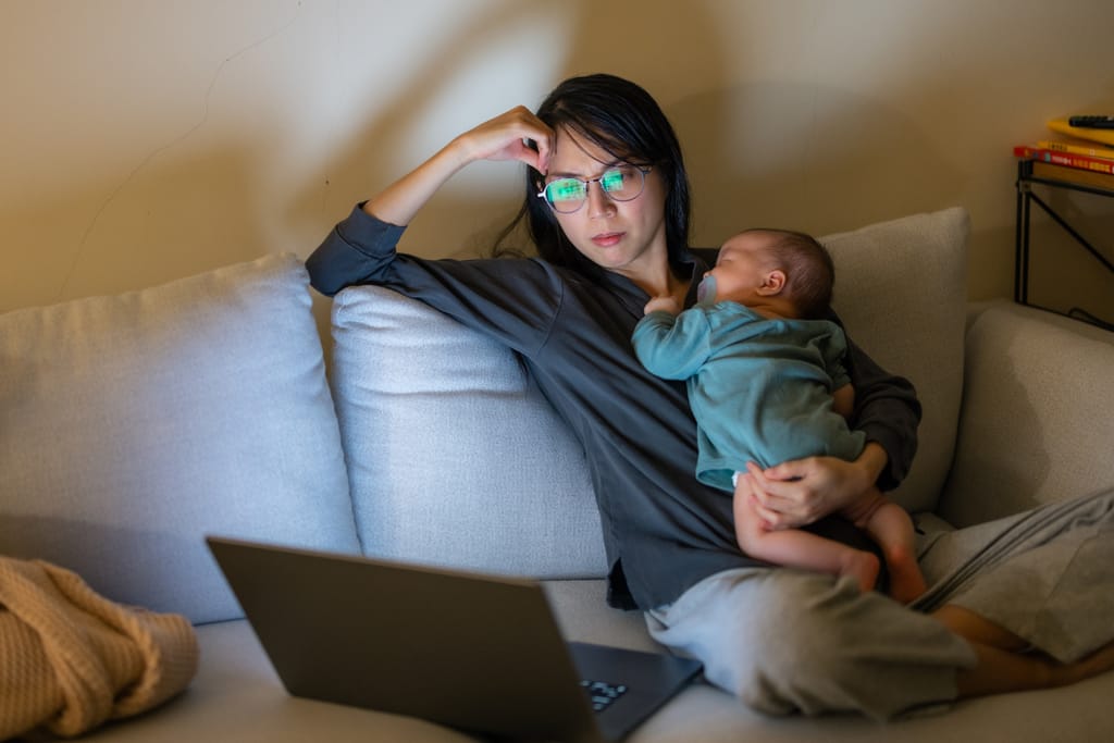 Äiti istuu sohvalla hämärässä olohuoneessa, pitää nukkuvaa vauvaa sylissään ja katsoo kannettavan tietokoneen näyttöä käsi otsalla, väsyneen ja keskittyneen näköisenä.