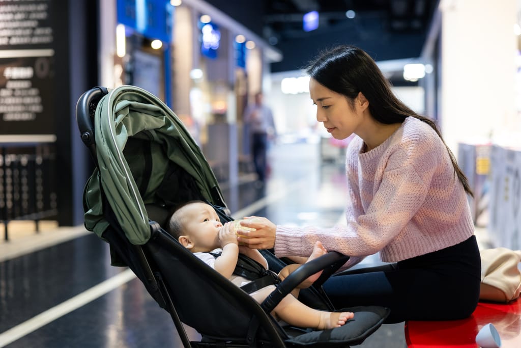 Nainen syöttää vauvaa tuttipullolla lastenrattaissa ostoskeskuksen käytävällä. Vauva makaa rattaissa ja katsoo naista, ympärillä näkyy valaistuja liiketiloja.