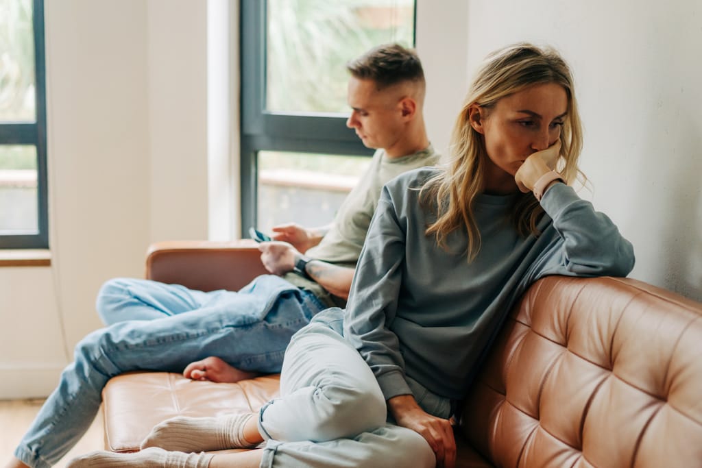 Pariskunta istuu erillään sohvalla; nainen katsoo mietteliäänä sivuun käsi suun edessä, kun mies selaa puhelinta taustalla.